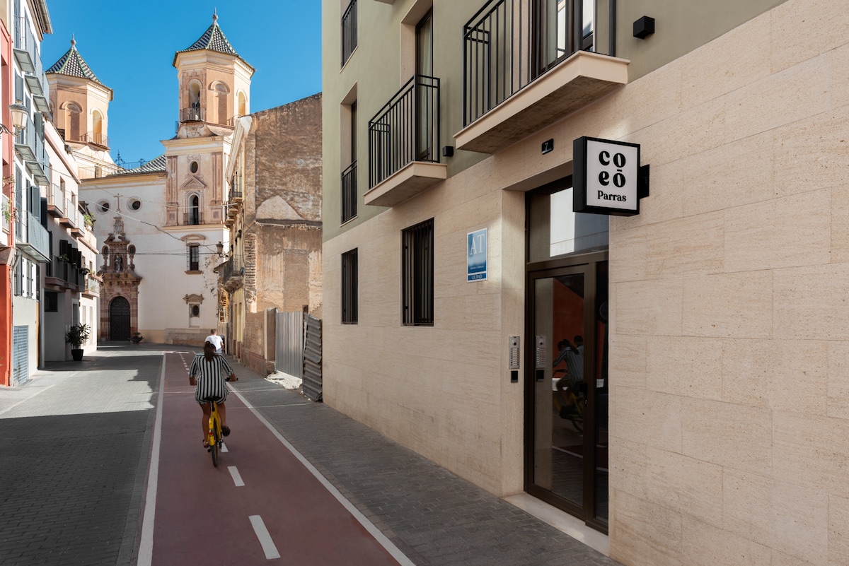 A view of the exterior of the Coeo apartments along a quiet street. Bicycles are parked nearby, and a cyclist is seen riding past. The building's inviting entrance sign, featuring 'Coeo Parras,' is visible. Historic architecture is present in the background.