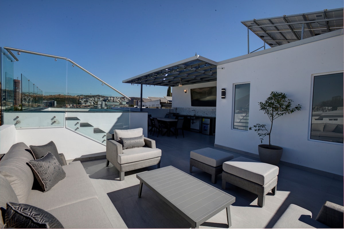 A rooftop terrace is presented with modern outdoor furniture, including a spacious sofa and lounge chairs, arranged around a sleek coffee table. A large shaded area with a dining table and a view of the surrounding landscape is also shown, providing a stylish space for gatherings.
