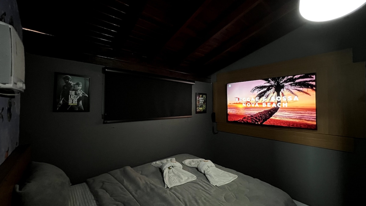 A dark-themed sleeping area features a spacious bed with neatly arranged towels. A large TV mounted on the wall displays a vibrant beach scene, while framed artwork adds character to the room. Soft lighting from a ceiling fixture creates a cozy ambiance.