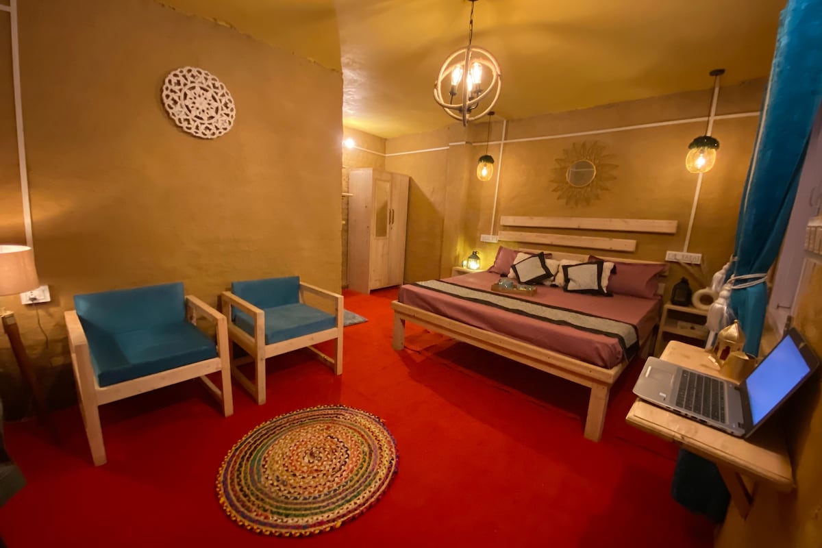 The room features mud plastered walls and a bright red carpet. A spacious double king-size bed is complemented by colorful cushions. Two blue chairs are positioned beside a round rug. A dedicated workspace with a laptop is visible, along with ambient lighting from hanging fixtures.