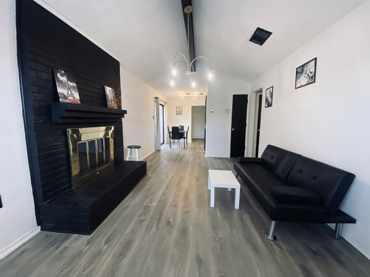 A living area is presented with a sleek black sofa and a small white coffee table. A dark, minimalist fireplace serves as a focal point against the contrasting light walls. Soft lighting is provided by a modern fixture overhead, creating a warm ambiance in this open space.