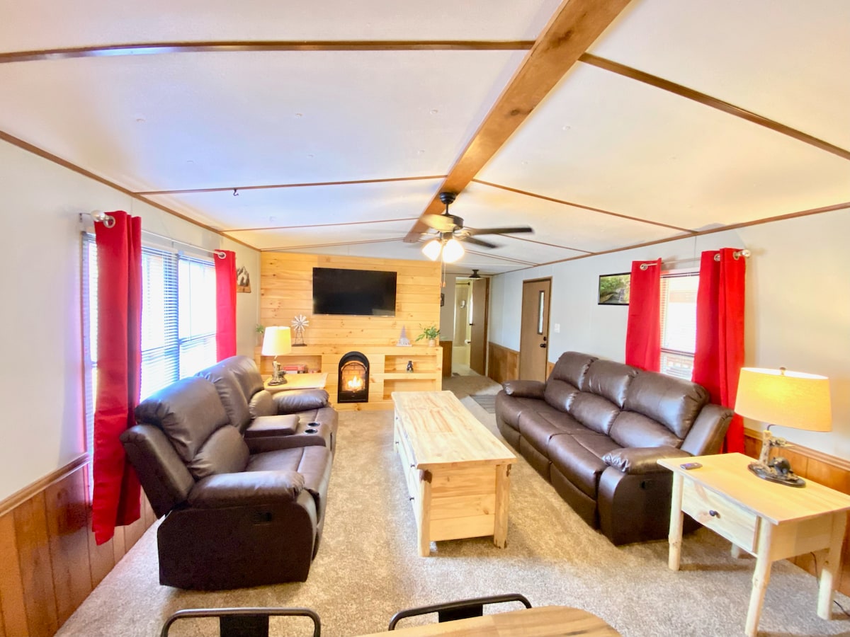 The family room features two dark leather sofas facing a central wooden coffee table. A large flat-screen TV is mounted on the wall above a cozy electric fireplace. Light streams in through windows adorned with red curtains, illuminating the room's warm, welcoming atmosphere.