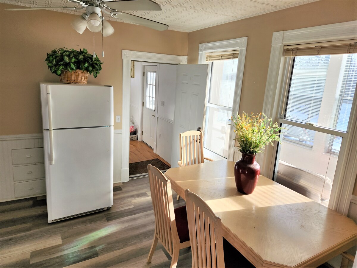 A bright and airy dining area features a wooden table surrounded by light wooden chairs. A vases with flowers is positioned at the center of the table. A white refrigerator is situated in the corner, with sunlight streaming in through large windows.