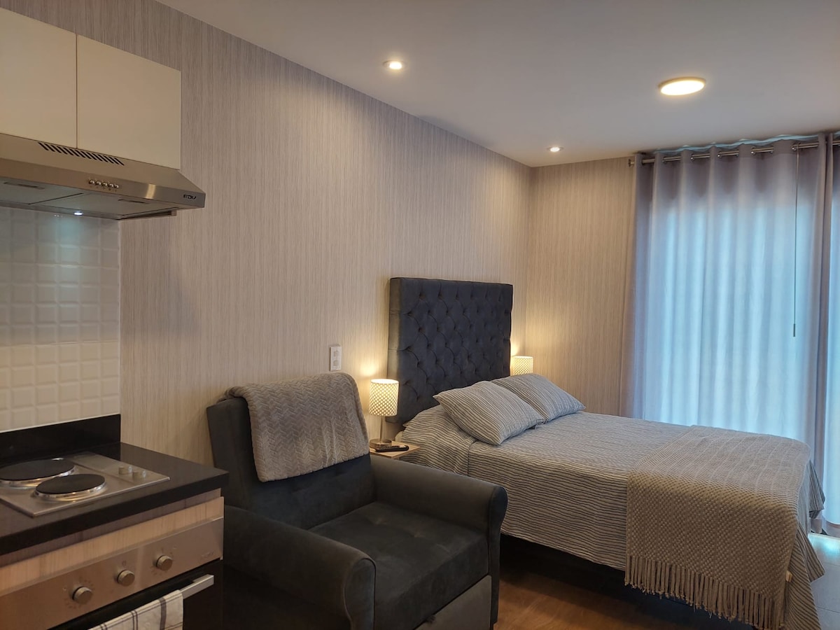 A cozy sleeping area is featured, with a spacious bed dressed in light bedding. A grey armchair sits nearby, and a small kitchen area is visible with a stovetop. Soft ambient lighting illuminates the room, complemented by large windows covered with sheer curtains.