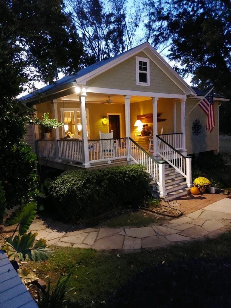 The exterior of a charming cottage is visible at dusk, featuring a covered porch with white railings. Potted plants and bright yellow mums are placed near the entrance. A warm light from inside casts a welcoming glow, and an American flag is displayed proudly.
