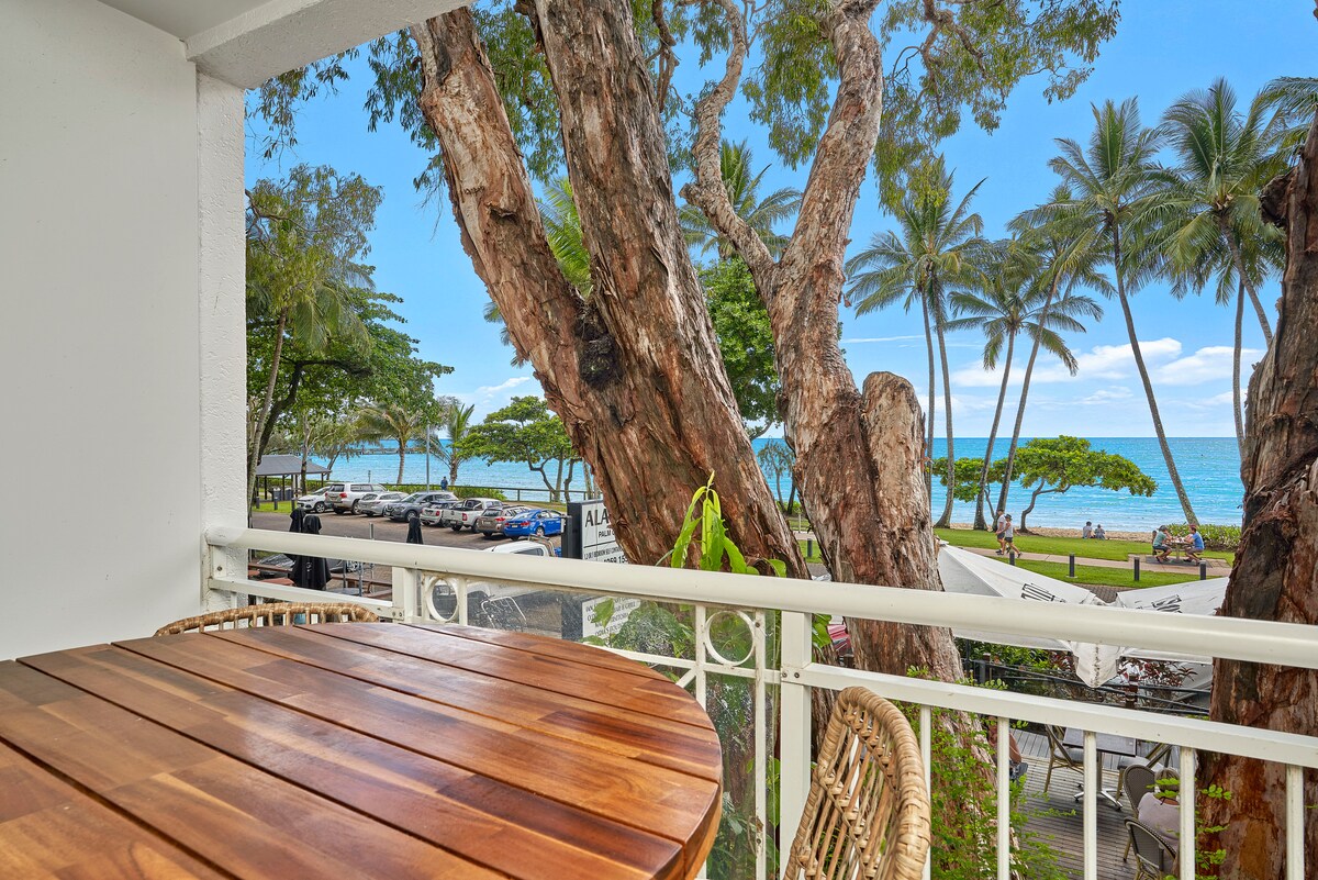 A balcony table, crafted from polished wood, is positioned for outdoor dining. The view showcases lush palm trees swaying gently and the sparkling Coral Sea in the distance, offering a serene setting, while nearby parked cars and greenery complement the vibrant atmosphere.