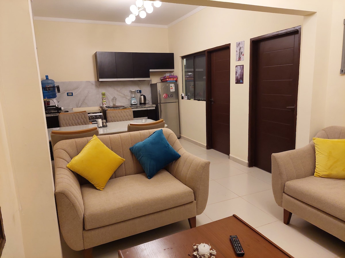 A well-lit living area features two comfortable sofas with yellow and blue accent pillows. A wooden coffee table sits in front, and the kitchen area is visible in the background, complete with modern appliances and cabinetry. Large windows allow natural light to fill the space.