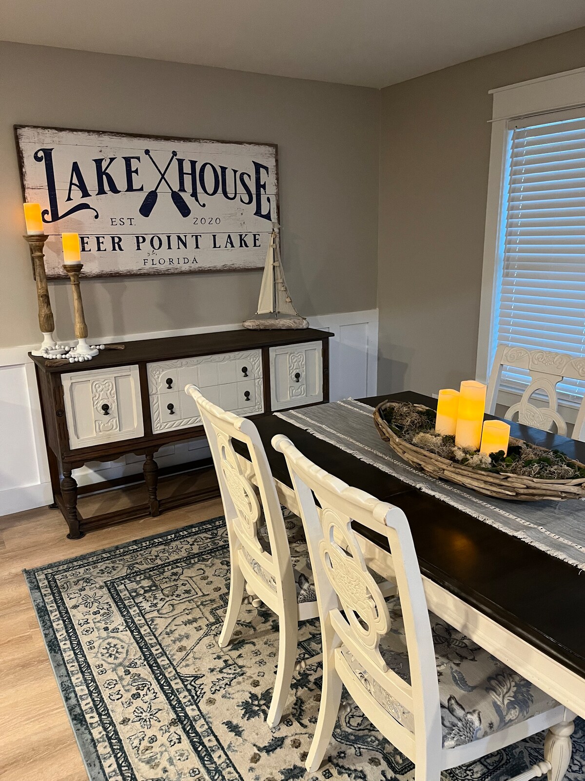A dining area is depicted, featuring a dark wooden table surrounded by white chairs. A decorative platter with candles is centered on the table. A sign reading 'Lakehouse' is displayed on the wall, complemented by warm candlelight and a cozy rug underfoot.