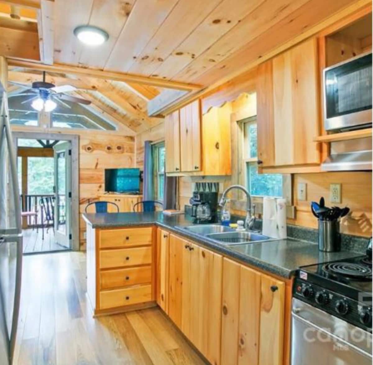 A rustic kitchen features wooden cabinets and countertops with a stainless steel sink. Modern appliances, including a stove, refrigerator, and coffee maker, are neatly arranged. Natural light enters through multiple windows, enhancing the warm wood tones throughout the space.