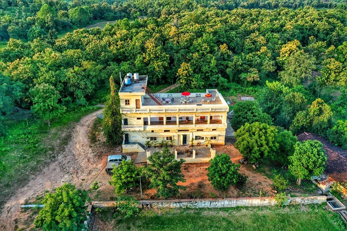 An aerial view captures a multi-level building surrounded by a dense green landscape. The structure features balconies and an outdoor seating area with red umbrellas, integrating harmoniously with the lush forest backdrop. Pathways and trees enhance the natural setting.