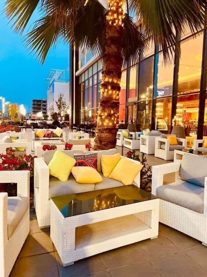 An outdoor seating area is presented with white wicker furniture, featuring plush cushions in shades of yellow and decorative accents. Potted flowers add color to the space, while a palm tree is wrapped in soft lighting, enhancing the evening ambiance.