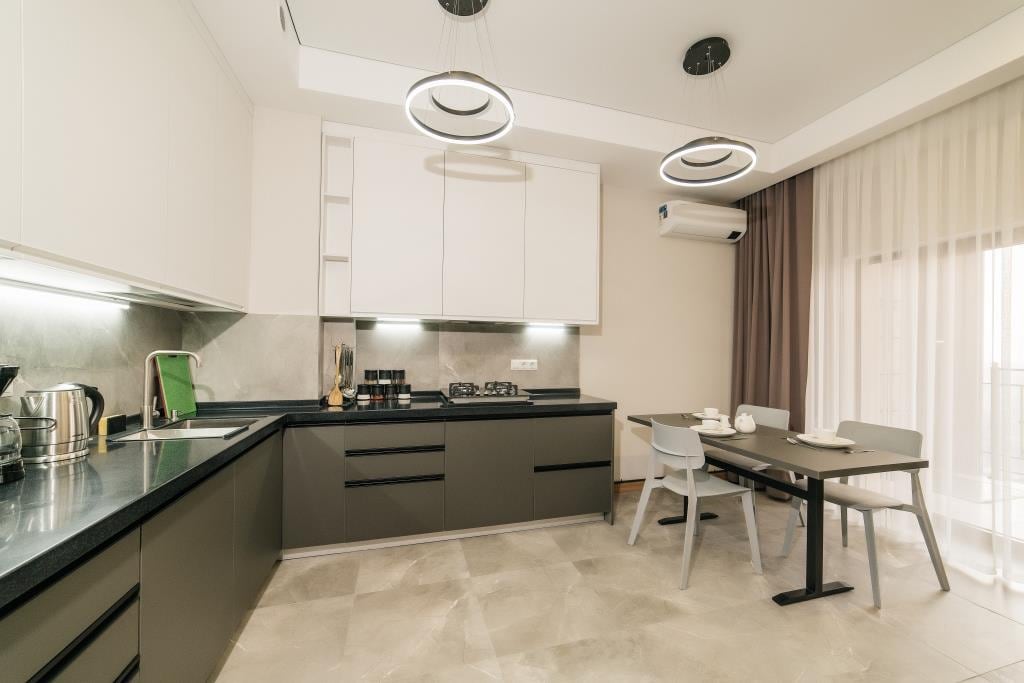 A modern kitchen features sleek cabinetry and a dark countertop, illuminated by circular pendant lights. A dining table with light grey chairs is positioned nearby, and large windows provide natural light, complemented by sheer curtains.