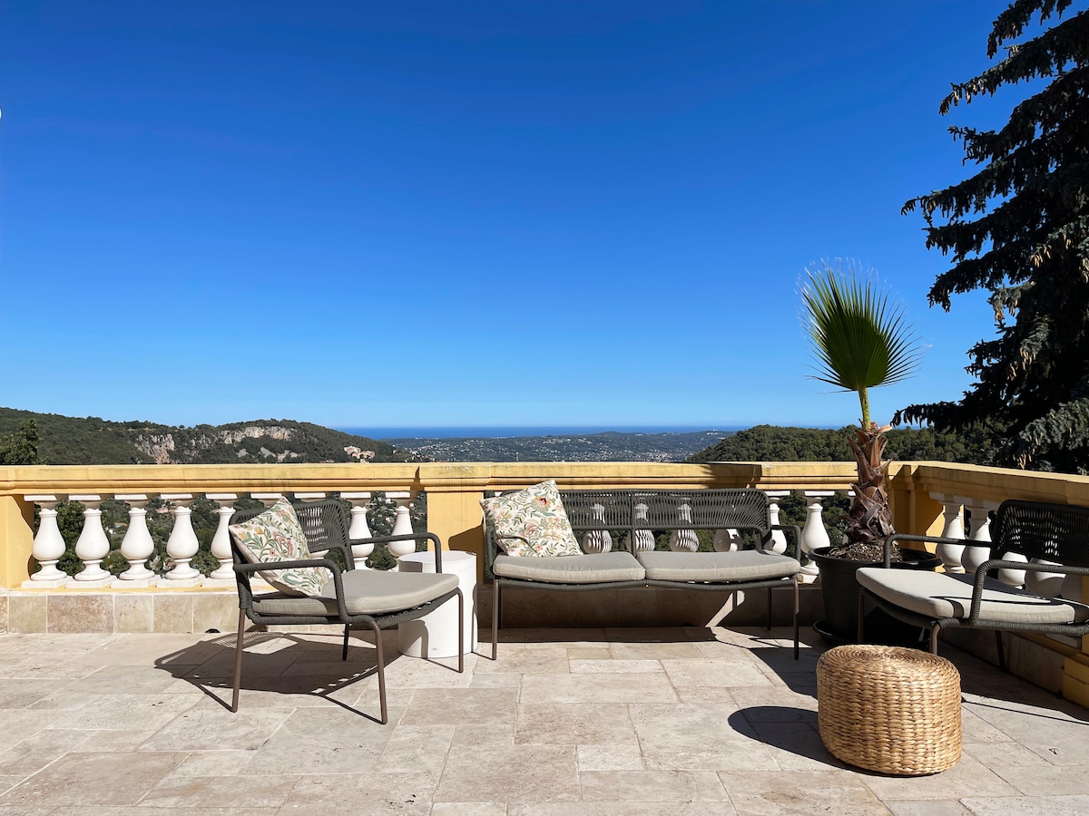An outdoor terrace is presented, featuring comfortable seating arrangements with plush cushions. A natural stone surface contributes to the elegant setting, while a view of rolling hills and the sea extends in the background under a clear blue sky.