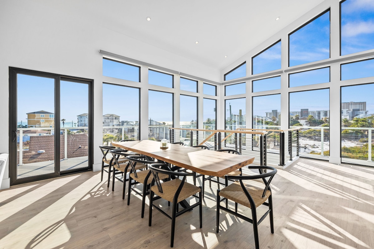 A spacious dining area features a long wooden table surrounded by stylish chairs. Floor-to-ceiling windows provide ample natural light and showcase views of the surrounding area, creating a bright and inviting atmosphere. The design incorporates clean lines and a modern aesthetic.