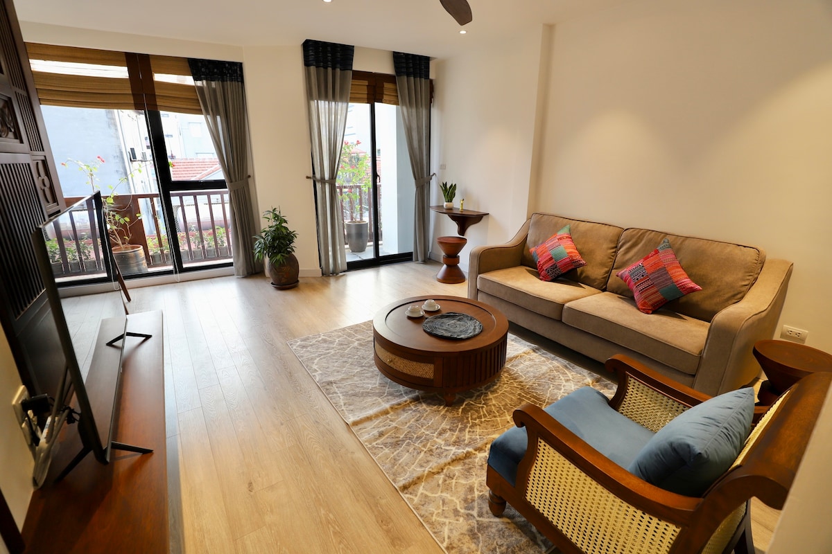 A bright living area features large windows allowing natural light to fill the space. A cozy sofa is complemented by colorful cushions, while a round coffee table sits on a textured area rug. Decorative plants add a touch of greenery to the inviting atmosphere.