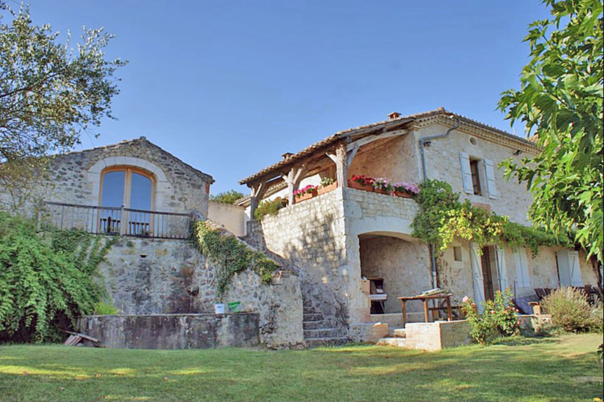 The exterior of a rustic country house showcases a stone structure with a wooden terrace adorned with flower pots. Steps lead from the garden to the main living area. A separate stone outbuilding is visible, nestled among greenery under a clear blue sky.