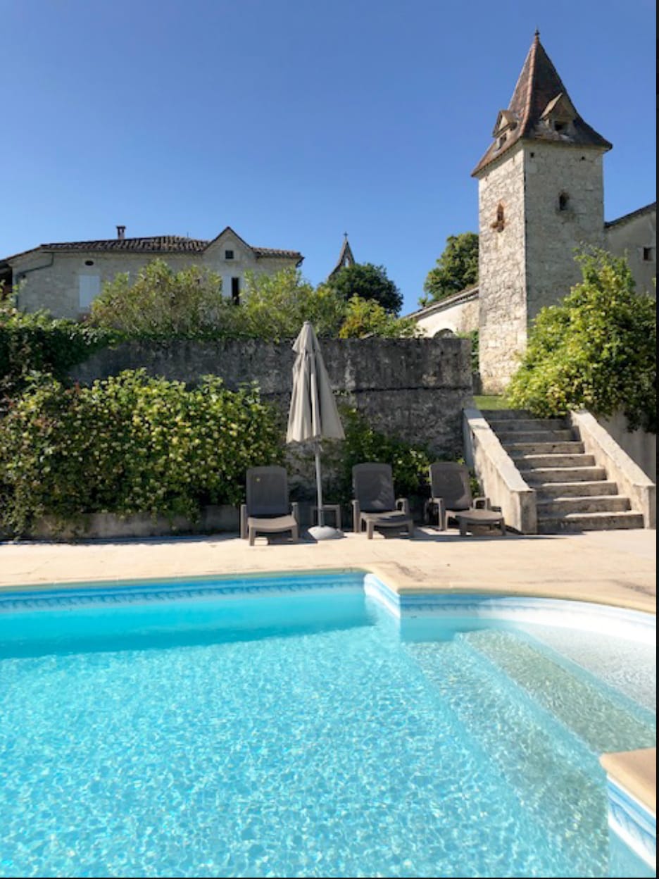 A private pool with clear blue water is framed by a spacious terrace featuring sun loungers. A stone structure with a tower rises in the background, surrounded by greenery and trees. Steps lead from the pool area to the garden above.