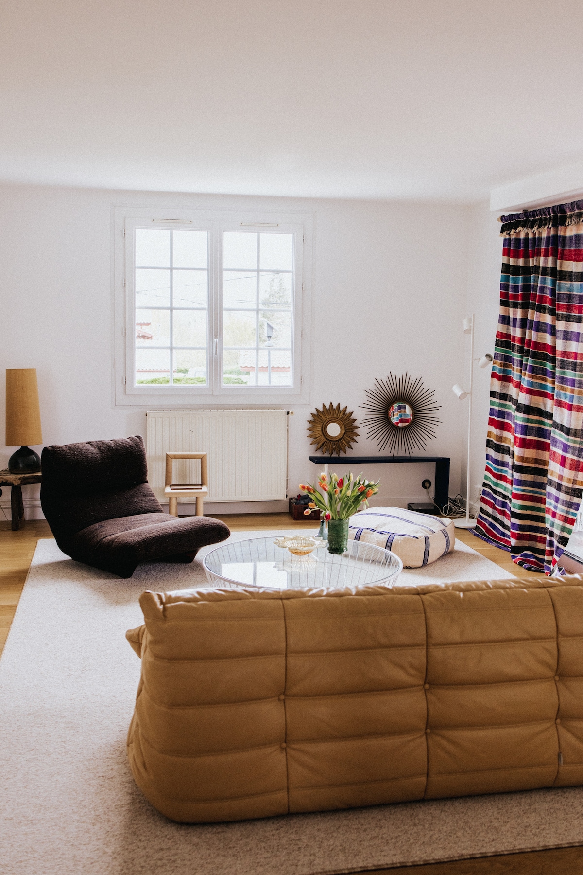 A bright living area features a mix of contemporary furniture, including two plush lounge chairs and a relaxed sofa. A round glass coffee table is centered on a neutral rug, with a vase of fresh flowers enhancing the space. Large windows allow natural light to fill the room.