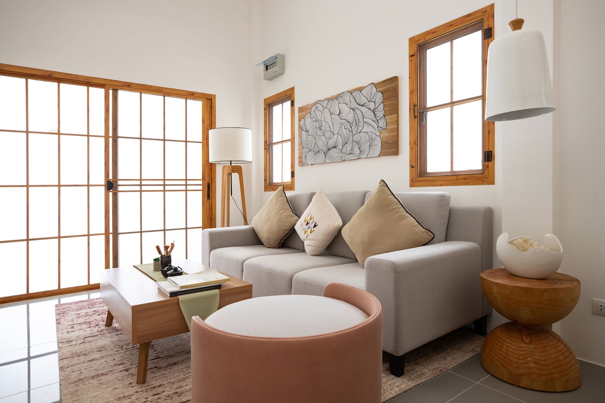 The living room features a light gray sofa adorned with decorative pillows, complemented by a wooden coffee table. Natural light enters through the large sliding doors and windows, while a contemporary lamp and a round accent table enhance the space.