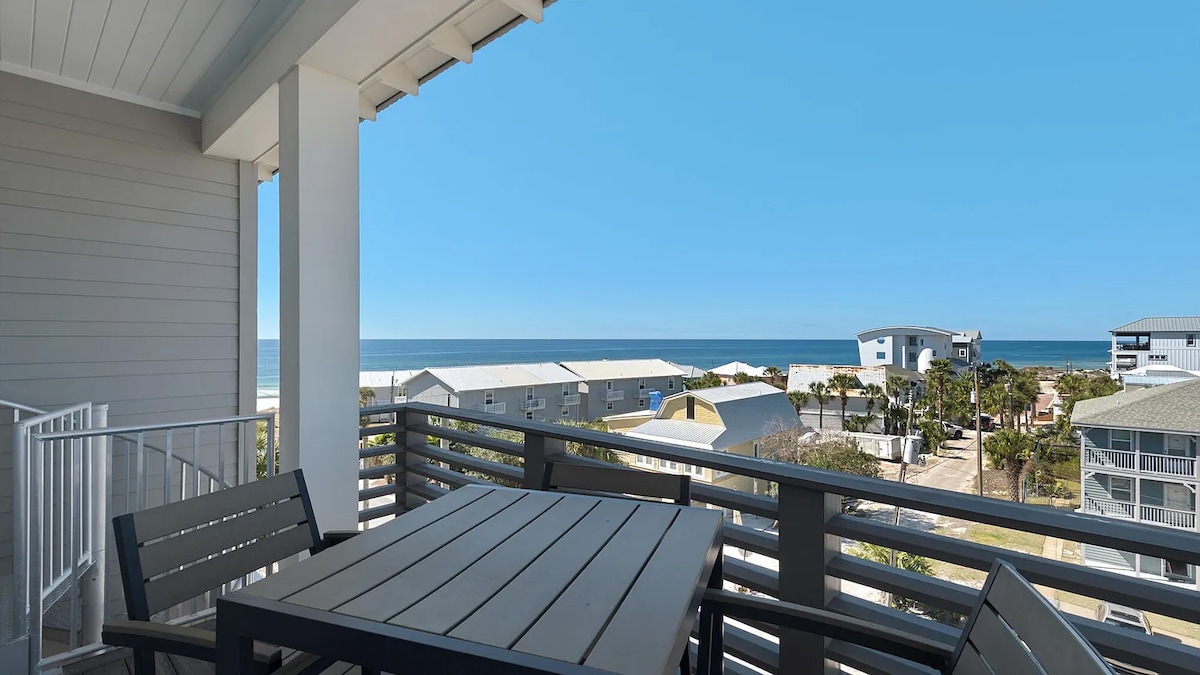A balcony view showcases the Gulf waters, with clear blue skies above. A wooden table and chairs are visible in the foreground, providing an inviting spot to enjoy the scenery. Surrounding buildings and palm trees offer a glimpse of the coastal environment.