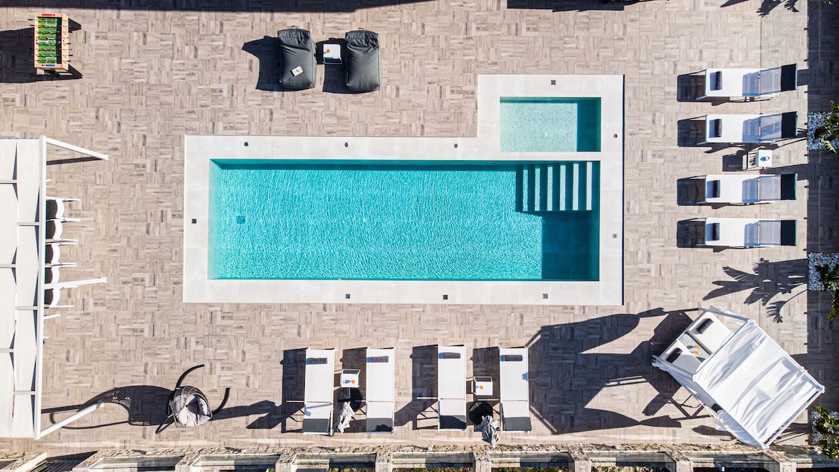 An expansive outdoor area features a large swimming pool surrounded by seating areas. Sun loungers are positioned along the poolside, with shaded spots and tables available for relaxation. A separate children's pool is visible, providing an inviting space for younger guests.
