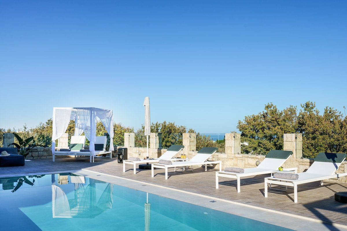 An outdoor area showcases a large swimming pool surrounded by several lounge chairs and sunbeds. A shaded cabana rests at one end, providing a cozy spot to relax. Lush greenery is visible in the background, with distant views of the sea.