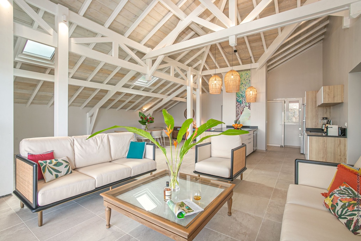 An airy living space is highlighted by high ceilings and a stunning exposed beam structure. Two light-colored sofas frame a central wooden coffee table, complemented by vibrant cushions. A well-equipped kitchen is visible in the background, with natural light entering through skylights.