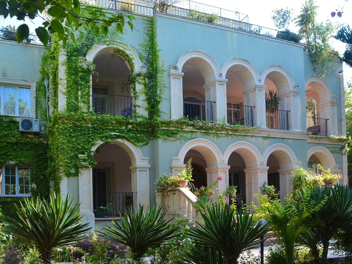 The exterior of a multi-story building is shown, featuring archways adorned with climbing plants. Balconies with wrought iron railings overlook a lush garden below, where a variety of tropical plants are visible. Sunlight filters through the greenery, creating a serene ambiance.