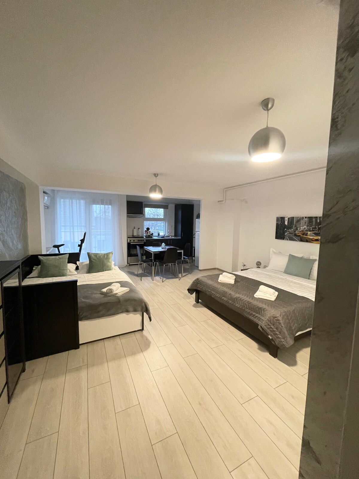 A spacious living area features two beds with neutral bedding, accompanied by a small dining table set for four. Natural light filters through large windows, while modern light fixtures overhead enhance the open layout. A kitchenette and lounge area are visible in the background.