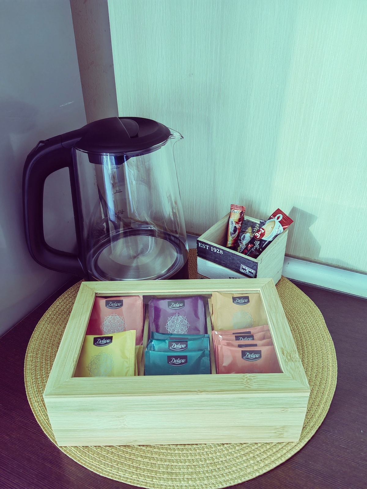 A kettle is placed beside a wooden box containing an assortment of tea sachets in various colors. A small container holds additional items, all situated on a textured mat, contributing to a welcoming refreshment area.
