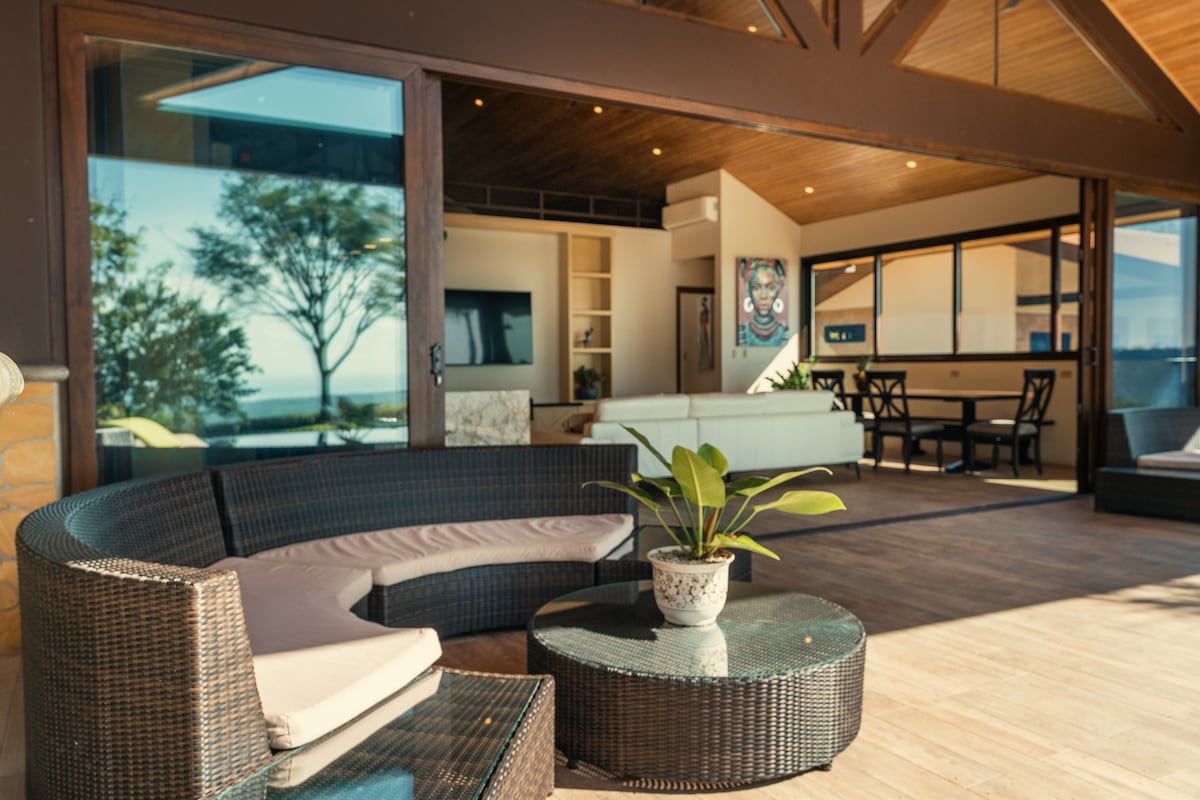 An inviting outdoor sitting area features a circular wicker sectional sofa, complemented by a round glass coffee table. Large sliding glass doors offer a view of the modern interior, while lush greenery and scenic ocean vistas are visible in the background.