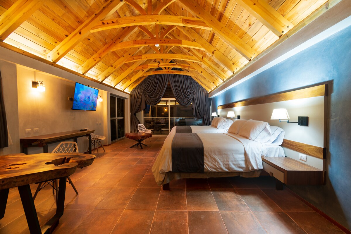 A spacious cabin-style suite features high wooden ceilings with exposed beams. Two large beds are positioned against the wall, complemented by bedside lamps. A flat-screen TV is mounted nearby, and large windows are dressed with dark curtains, allowing natural light to fill the room.