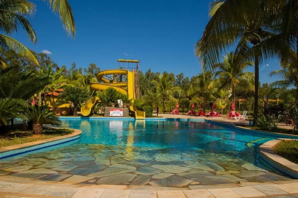 A large swimming pool is surrounded by palm trees and features a vibrant yellow waterslide in the background. Laid-back lounge chairs are positioned on the pool deck, inviting relaxation. The clear blue sky reflects off the tranquil water's surface, creating a serene environment.
