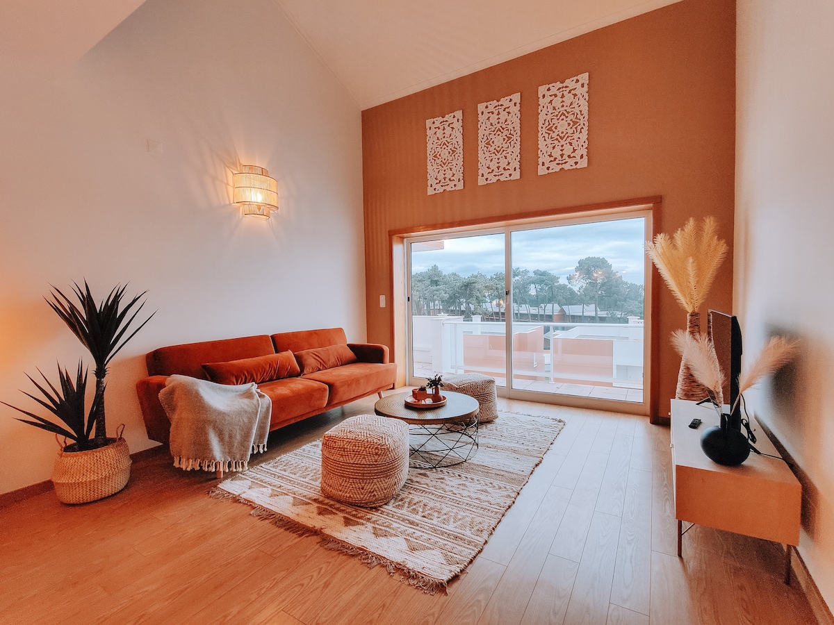 A spacious living area presents a warm sofa in rich tones, accompanied by decorative cushions. Natural light floods in through large windows, revealing outdoor views. A circular coffee table is centered on a textured rug, while potted plants add a touch of greenery to the room.