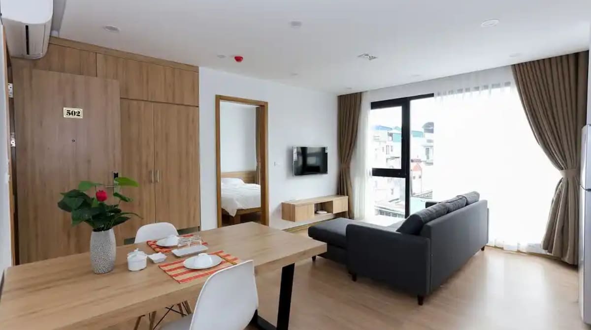 The open living area features a comfortable sofa positioned near a large window, allowing ample natural light. A dining table is set with tableware, complementing wooden cabinetry and a modern television mounted on the wall. Light-colored curtains frame the window, enhancing the space's brightness.