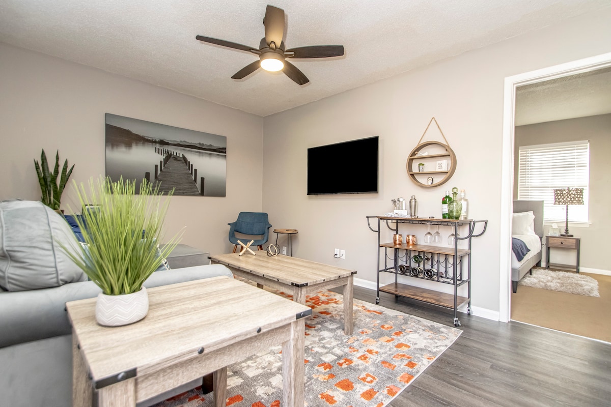The living area features a soft blue couch and an armchair, paired with two wooden coffee tables. A 42-inch smart TV is mounted on the wall opposite, while a bar cart with beverages is visible beside it. Light colored walls enhance the spacious feel.