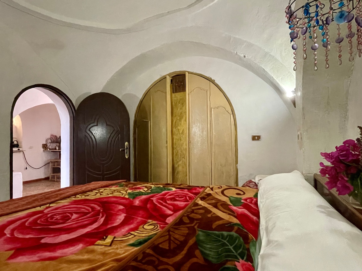 A cozy bedroom features an ornate bed covered with a vibrant floral blanket. Arched ceilings and a wooden wardrobe contribute to the room's charm. A small seating area is visible in the background, softly illuminated by wall sconces, creating a warm ambiance.