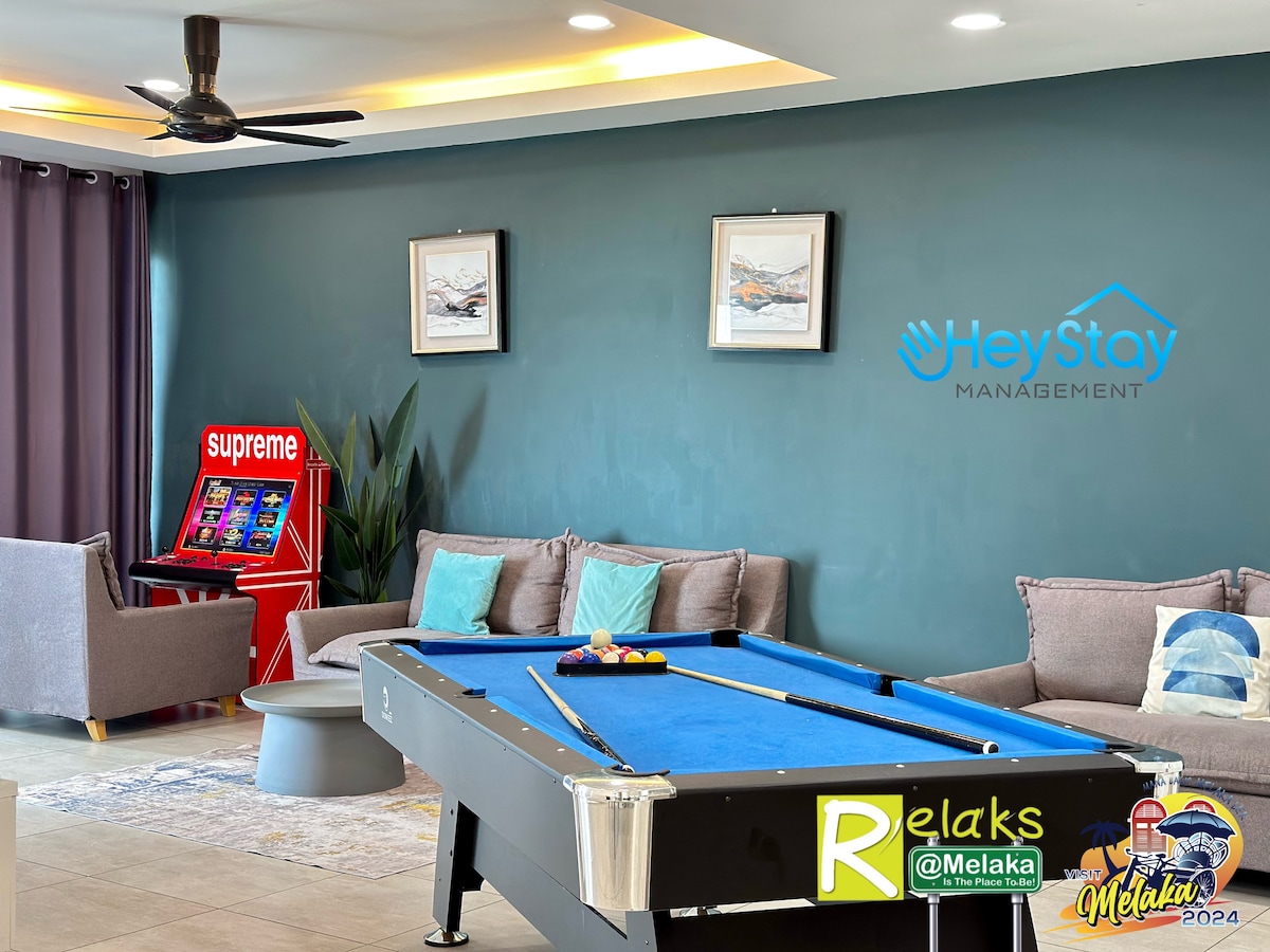 A spacious recreational area is showcased, featuring a blue pool table at the center. Comfortable seating is arranged nearby, complemented by wall art and a gaming machine. Soft natural light enters from large windows, while the walls are painted in a calming shade of green.