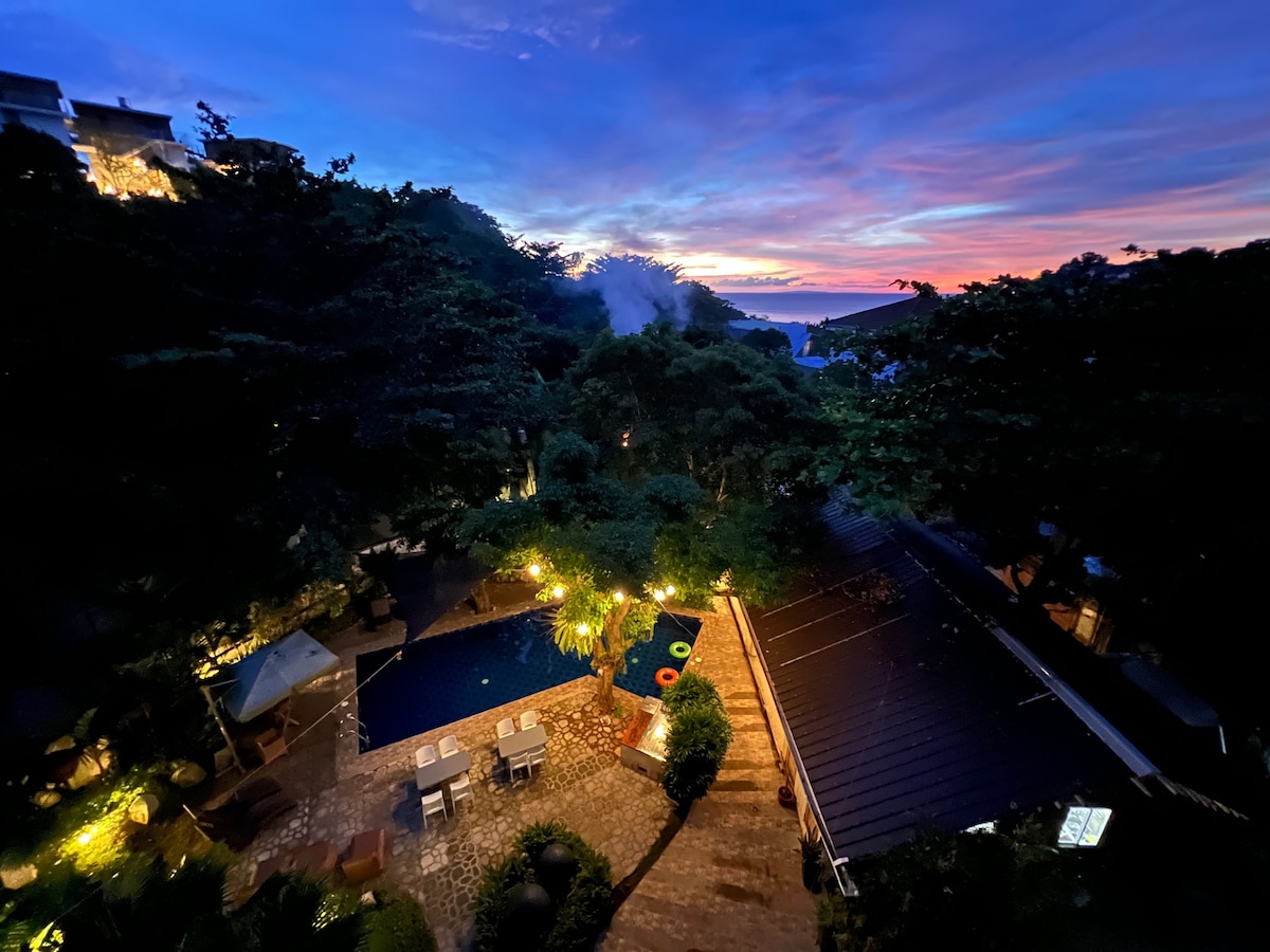 An expansive view reveals a swimming pool surrounded by lush greenery, illuminated by soft outdoor lighting. The sky transitions from deep blue to soft hues of orange and pink, hinting at a picturesque sunset over the horizon.