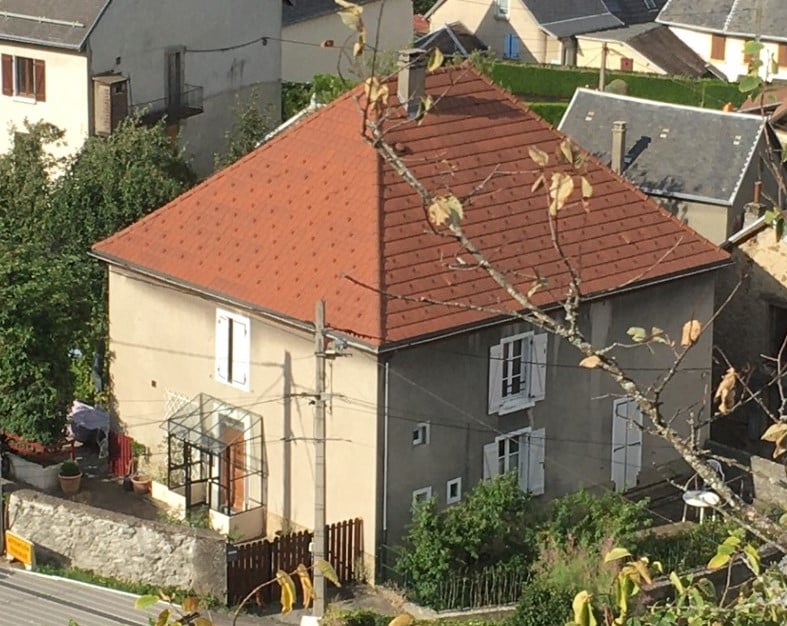A two-story house is visible, featuring a distinct red tile roof and white window shutters. The exterior is painted in neutral tones, with a small garden visible in front. Surrounding vegetation and other buildings provide a scenic backdrop.