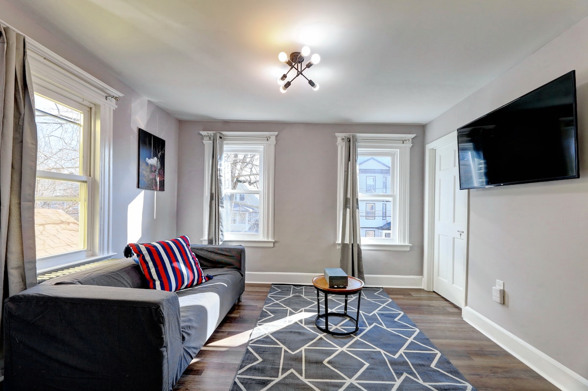 A bright living room features a comfortable couch with striped cushions. Two windows provide natural light, while a round coffee table is placed on a patterned rug. A wall-mounted television is visible, enhancing the space suited for relaxation.