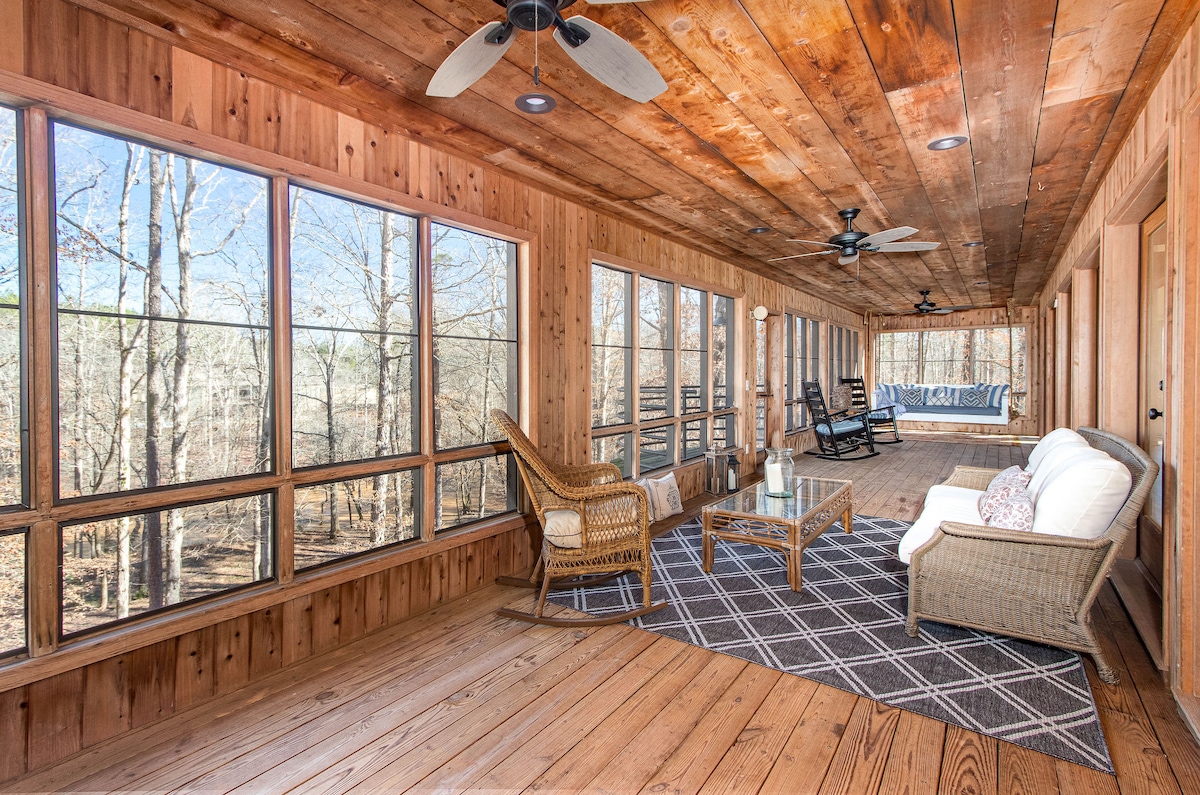 The screened-in porch features wooden walls and a rich wooden ceiling, providing a rustic ambiance. Comfortable seating includes a soft couch, wicker chairs, and a coffee table. Large windows allow natural light to fill the space and offer views of the surrounding trees.