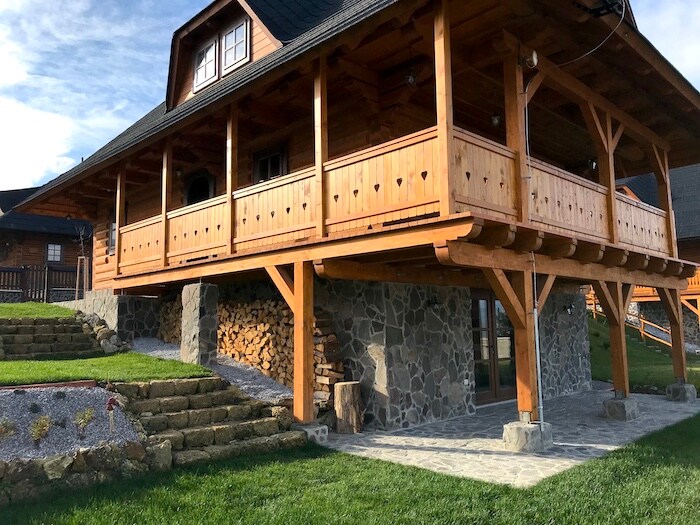 A charming wooden cottage is featured with a spacious raised terrace supported by stone columns. The exterior showcases a combination of wood and stone, with neatly stacked firewood beside the entrance. Green grass surrounds the building, enhancing the natural setting.