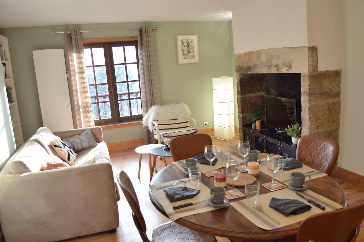 A living area features a comfortable couch and a round dining table set for six, with plates and glasses arranged neatly. Natural light enters through a large window framed with light curtains, illuminating the stone fireplace beside a TV stand and decorative elements.