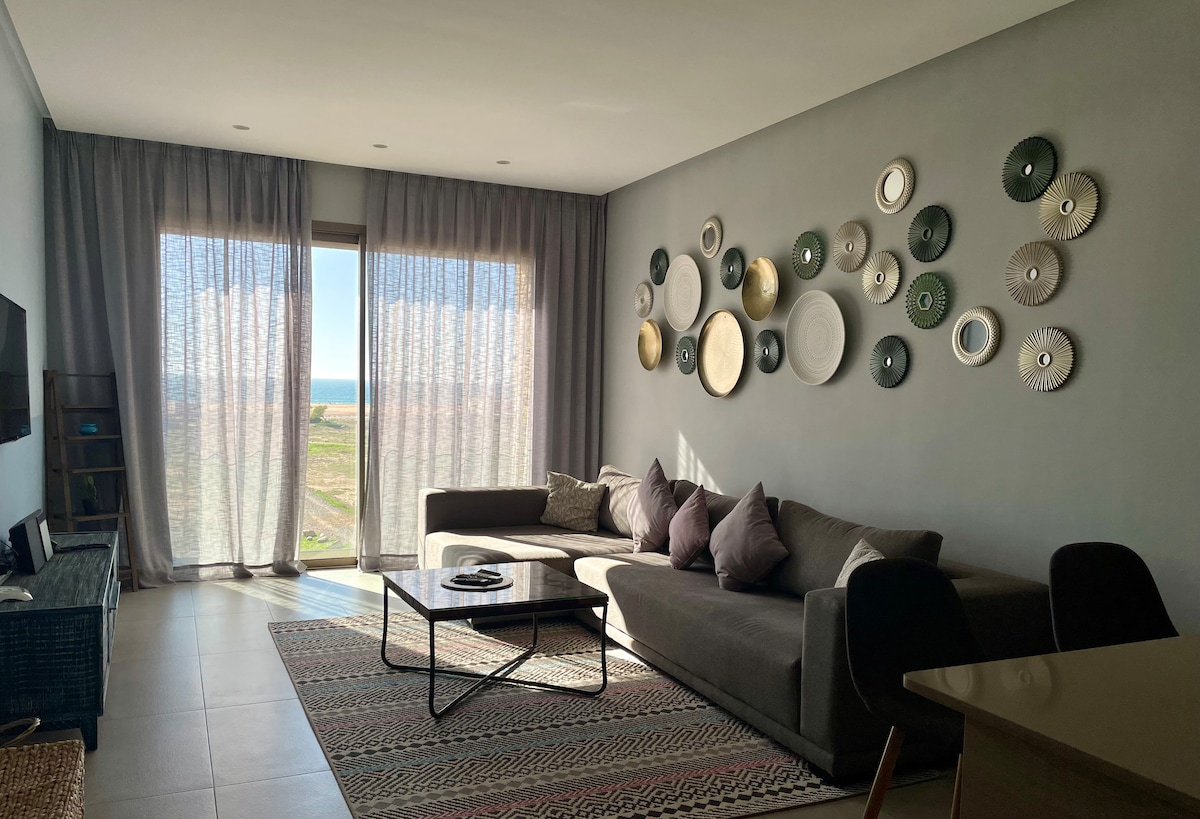 A comfortable living area showcases a dark gray sofa adorned with soft cushions. A coffee table rests on a textured rug, while decorative plates are arranged artistically on the wall. Natural light flows through large windows with sheer curtains, offering views of the sea.
