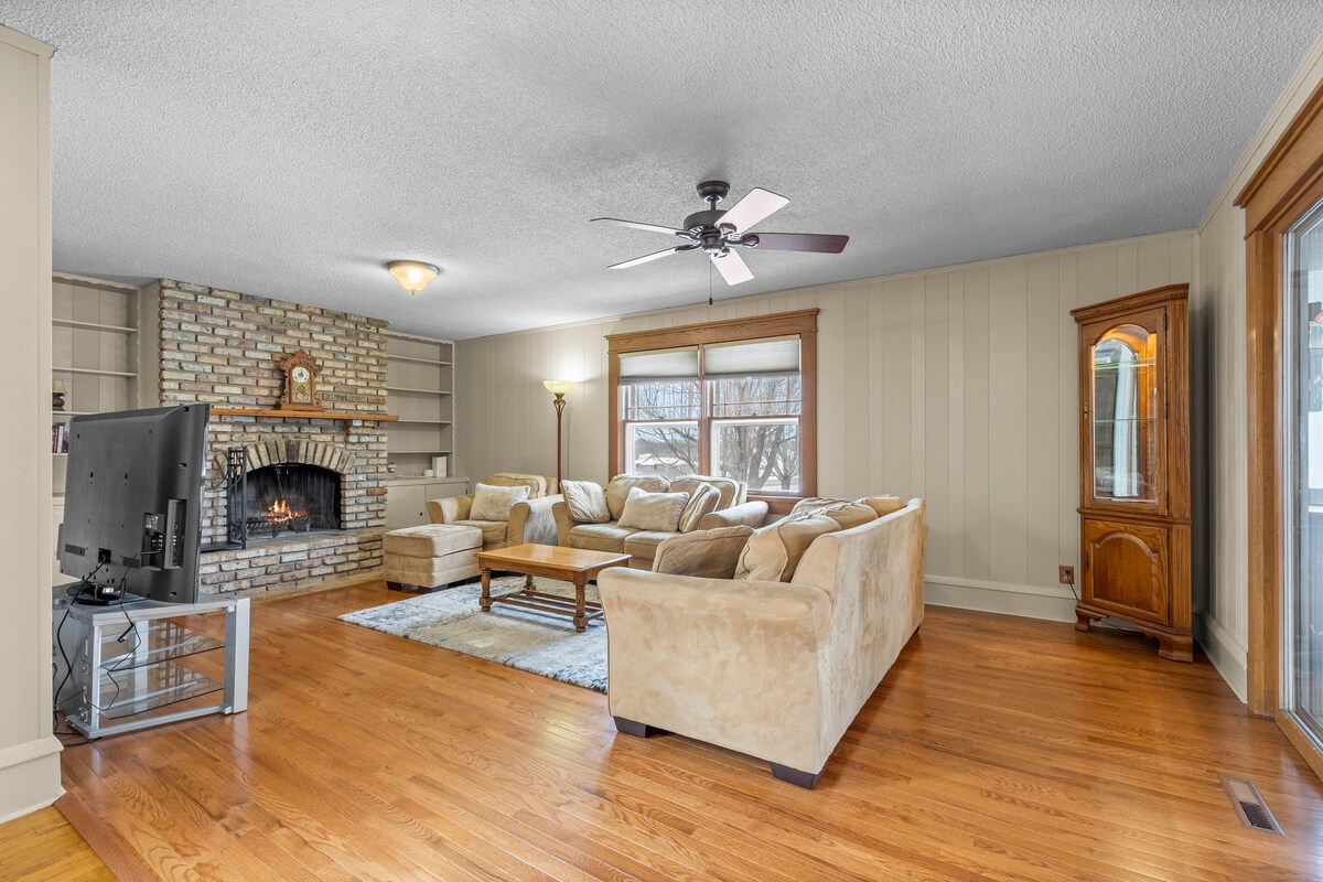 A spacious den features a stone fireplace with a wooden mantle and a ceiling fan. Comfortable couches are arranged around a central coffee table, creating an inviting gathering space. Natural light filters in through multiple windows, illuminating the hardwood floors.
