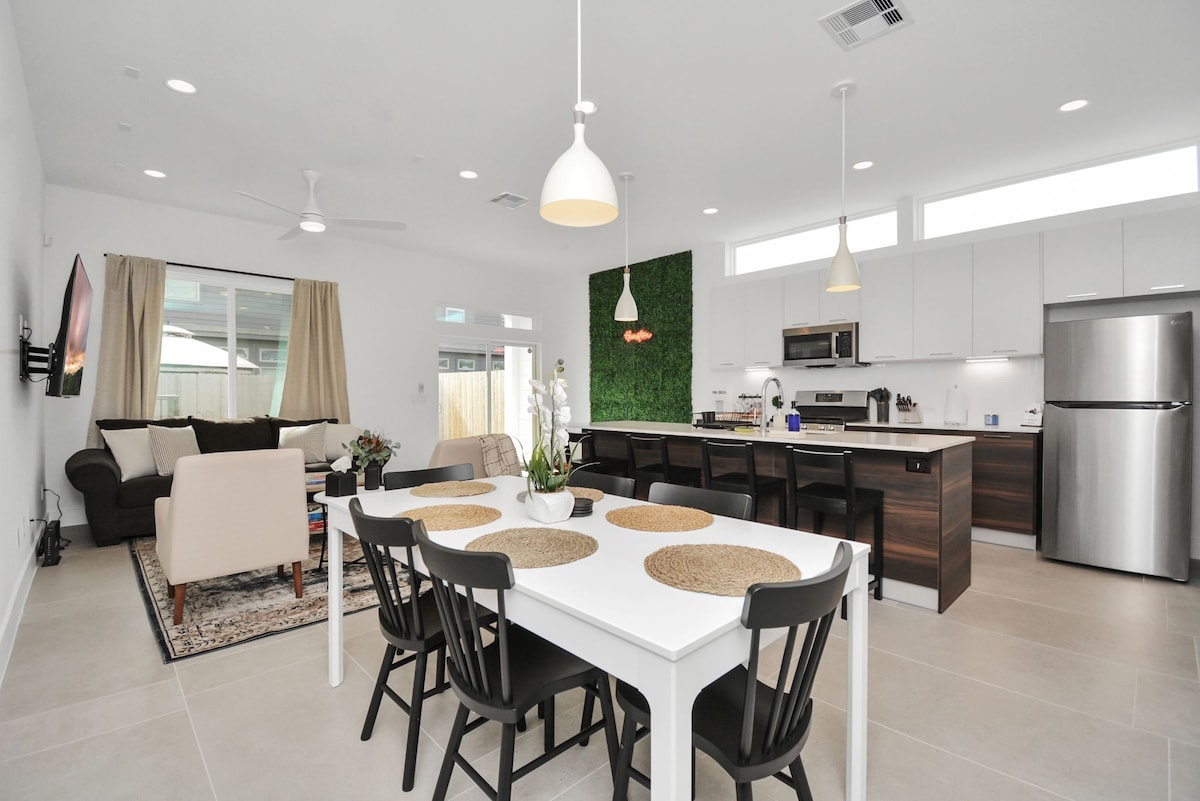 An open-plan living and dining area features modern furnishings, including a sofa and armchairs. A dining table with black chairs is set against a light backdrop, while a fully equipped kitchen is visible. Natural light fills the space through large windows and subtle greenery is displayed on a wall.