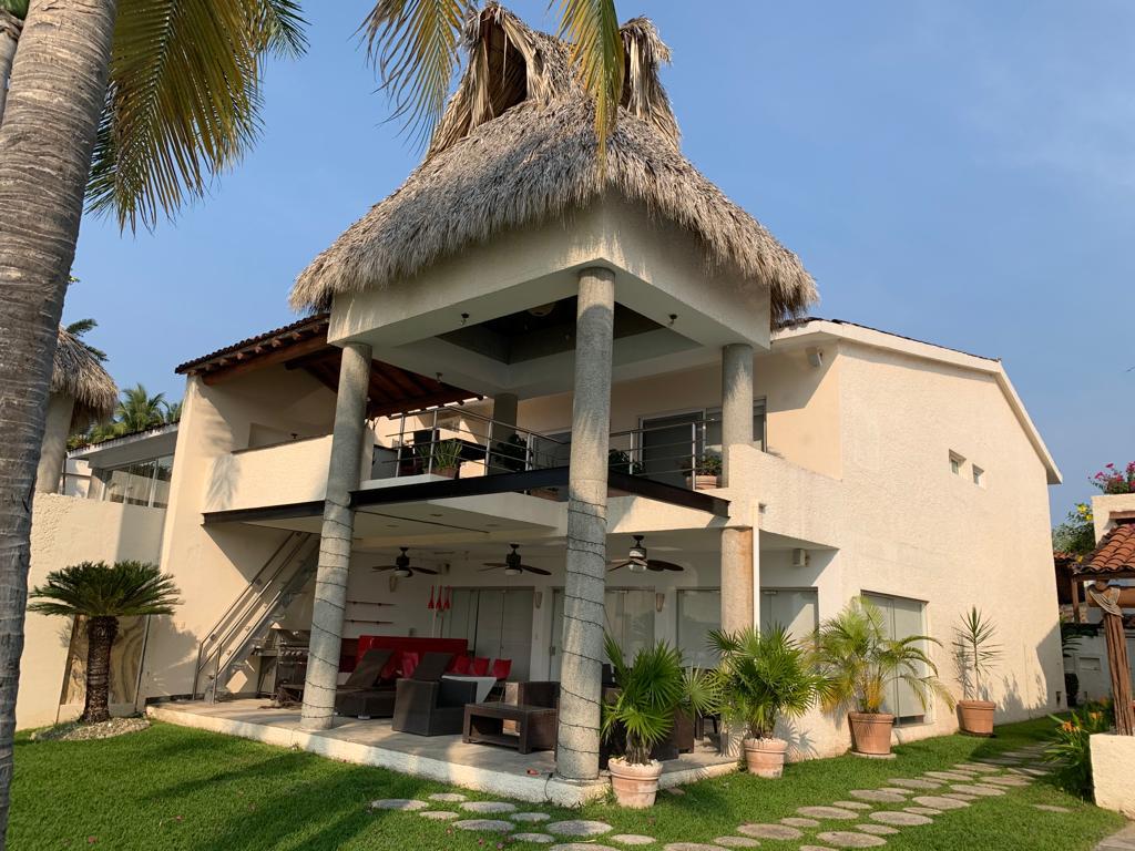 A two-story villa is observed, featuring a thatched roof and open terraces. Vertical columns support the upper level, which includes outdoor seating. Lush green grass surrounds the structure, and potted plants add warmth to the outdoor area. The overall design integrates natural elements with modern architecture.