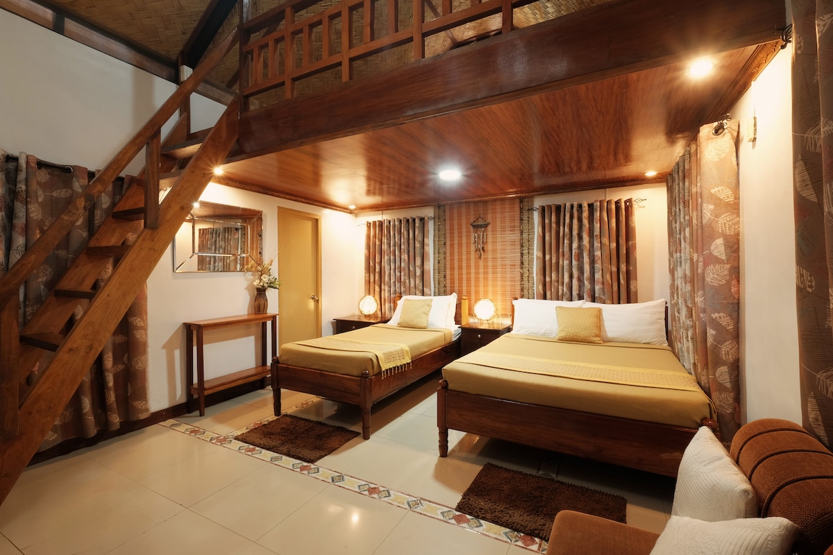 The room features two spacious beds and a cozy seating area with a brown sofa. A wooden staircase leads to the loft, where additional sleeping space is available. Natural light filters through the curtains, enhancing the warm wood finishes throughout the room.
