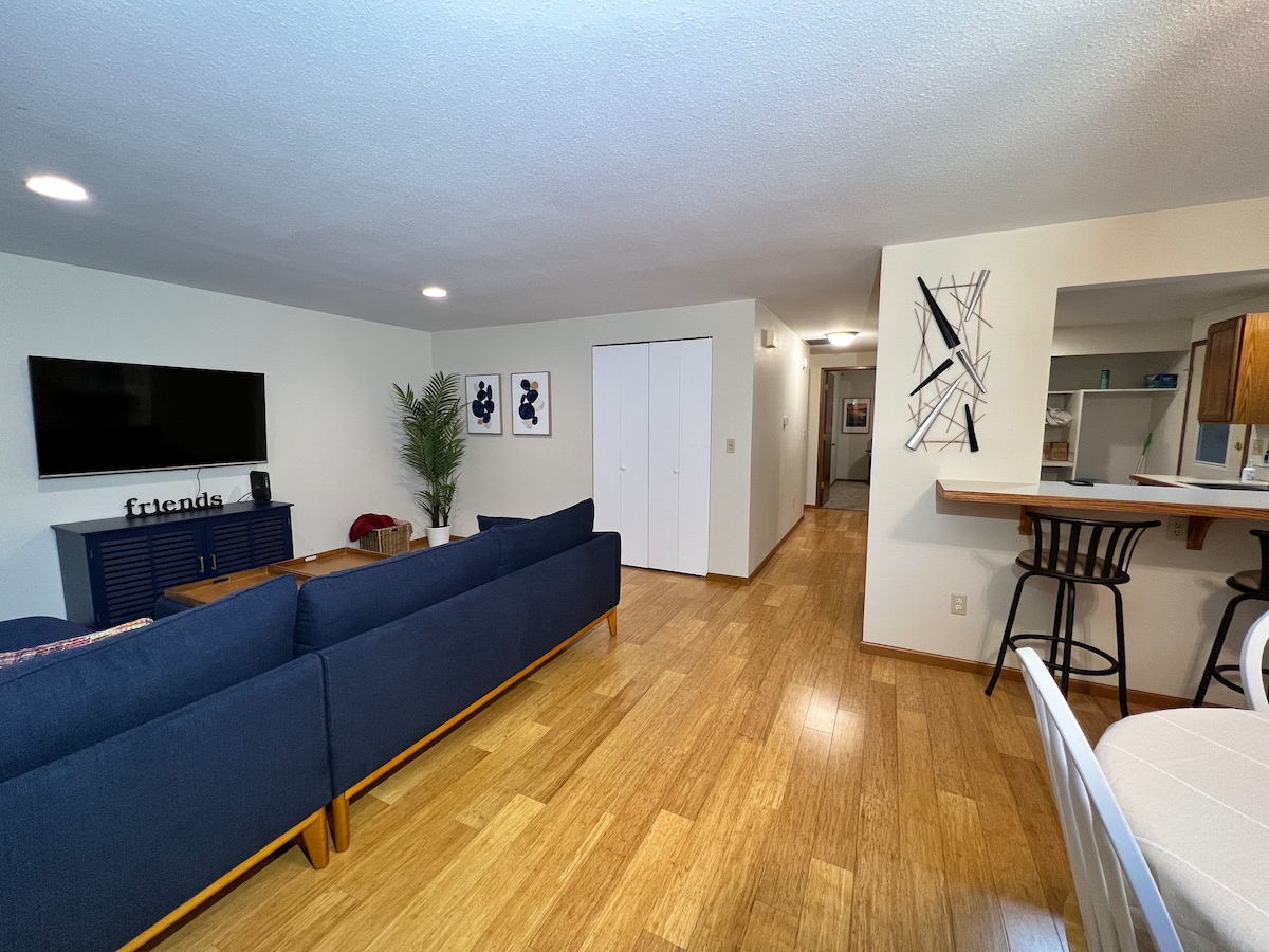 A spacious living area is furnished with a navy blue couch and a wall-mounted television. Light wood flooring complements the neutral wall color. A hallway leads to other areas of the house, while a dining area with bar seating is visible in the background.